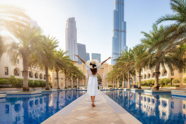 A woman in a white dress with arms raised stands by a pool with Dubai skyscrapers in the background.