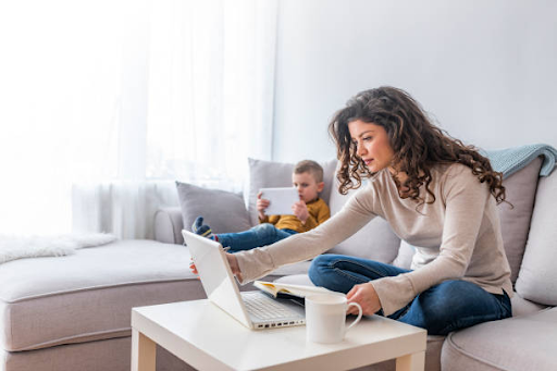 A freelancer working on a laptop at home, with a child playing in the background, relating to Dubai visa costs.